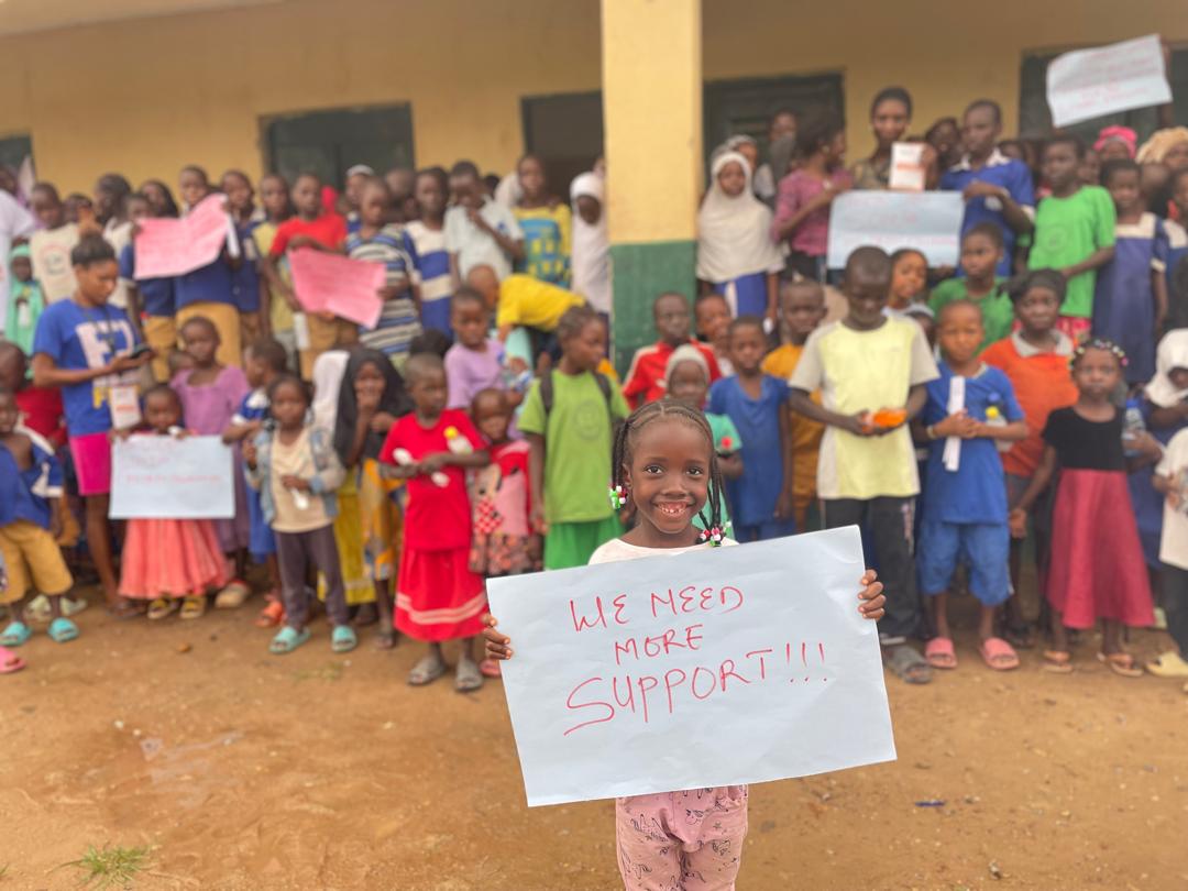 A child holding a sign that reads WE NEED MORE SUPPORT!!!